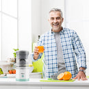 Hombre en cocina sosteniendo vaso de jugo junto a exprimidor eléctrico de frutas portátil con naranjas cortadas